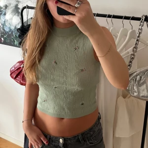 Ljusgrön stickad ärmlös topp - Supersöt ljusgrön stickad topp med höghalsad krage och croppad passform. Toppen har små broderade blommor i rosa och vitt samt fina hålmönster längs hela plagget. 