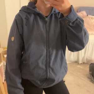 Fjällräven jacka - Säljer en blå vindjacka från Fjällräven, modell Nordli Jacket. Jackan har huva med snörning, dragkedja framtill och klassisk Fjällräven-logga på ärmen. Materialet är slitstark bomull och polyesterm. Storlek S 