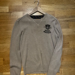 Beige stickad tröja från Morris - Snygg beige stickad tröja från Morris med broderad logga och emblem på bröstet. Tröjan har rund halsringning, ribbade muddar och lång ärm. Perfekt för dig som gillar klassisk och stilren look.