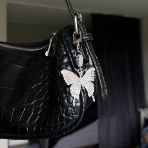 Väsksmycke/Nyckelring (fjäril) - Snyggt hängsmycke i form av en fjäril, täckt med glittrande stenar på framsidan. Perfekt att fästa på väskan eller som nyckelring. Ger en extra touch av bling och är superfin som accessoar. Önskar du veta exakta mått? Tveka inte att höra av dig!