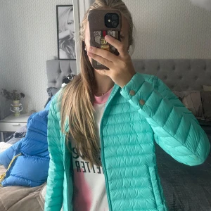 Turkos pufferjacka med dragkedja - Säljer en snygg turkos pufferjacka med dragkedja framtill och tryckknappar vid ärmsluten. Jackan är lätt vadderad och har en modern, sportig look som passar perfekt till kyligare dagar. Enkel att matcha med olika outfits.