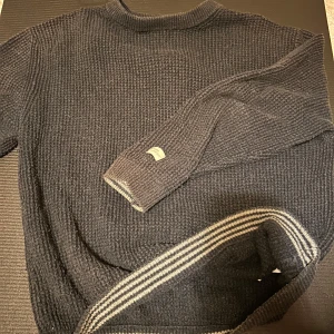 Mörkgrå stickad tröja från Zara - Mysig mörkgrå stickad tröja från Zara med ribbad krage och ärmslut. Snygga vita ränder vid halsen och nederkanten ger en sportig touch. Perfekt för kyliga dagar och enkel att matcha med jeans eller kjol. Funkar i många storlekar 