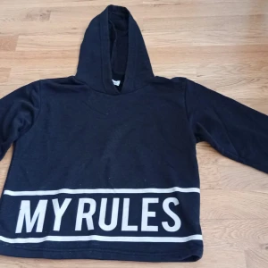 Svart hoodie med text MY RULES - Svart hoodie från Atababy med stor vit text MY RULES längs nederkanten. Tröjan har huva, långa ärmar och en loose passform. Perfekt för dig som gillar en chill och självsäker stil.