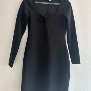 Svart klänning med knyt från H&M Divided - Svart, stilren kortklänning från H&M Divided med långärmad design och fyrkantig urringning. Snygg detalj med knyt framtill. Klänningen har en figurnära passform och är gjord i ett mjukt syntetmaterial som sitter bekvämt mot kroppen.