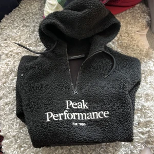 Svart fleece hoodie Peak Performance - Mysig svart hoodie från Peak Performance i fleece med broderad logga framtill. Tröjan har huva med snörning och half zip-dragkedja. Perfekt för kyliga dagar och riktigt skön att dra på sig när man vill vara bekväm.