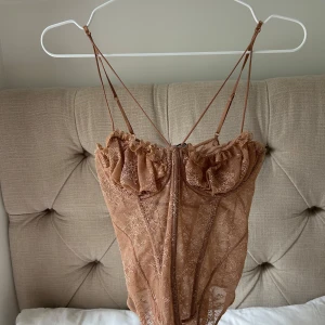 Brun spetsig korsettopp från Zara - Supersnygg brun korsettopp från Zara med tunna axelband och transparent spetsmaterial. Toppen har volangdetaljer vid bysten och en figurnära passform som ger en trendig look. Perfekt att styla med jeans eller kjol för en cool vibe.