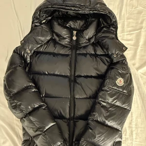 Svart Moncler dunjacka för DAM - Säljer en svart, glansig dunjacka från Moncler med huva och dragkedja framtill. Jackan har klassisk pufferstil, logotyp på ärmen och snygga detaljer. Perfekt för kalla dagar när du vill hålla stilen och värmen. Om någon vill ha bild med jackan på så kan ni skriva.