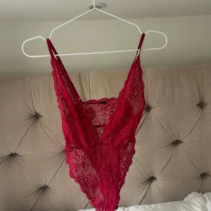 Röd spetsbody med djup v-ringning - Säljer en röd body i spets med djup v-ringning och tunna axelband. Litet hål/slitning på sidan.