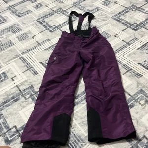 Lila täckbyxor med hängslen stl 134/140 - Säljer ett par lila täckbyxor med svarta förstärkningar vid bensluten och praktiska hängslen. Byxorna har dragkedja och knapp framtill samt en ficka med dragkedja på benet. Perfekta för vinterns alla äventyr.