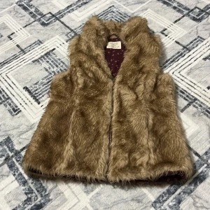 Beige pälsväst från Zara Girls - Supermjuk och fluffig beige pälsväst från Zara Girls. Västen har en klassisk rak passform och är fodrad med ett vinrött tyg med små vita blommor. Perfekt att slänga över en hoodie eller långärmad tröja för en cool lager-på-lager-look.