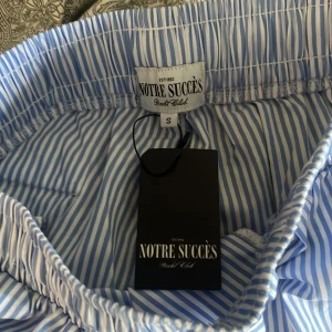 YACHT CLUB STRIPED PANTS från Notre Succès S - Snygga blå- och vitrandiga byxor från Notre Succès Yacht Club. Byxorna har rak passform, elastisk midja och snörning framtill. Perfekta för en chill och stilren look.