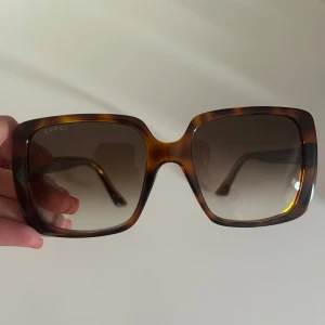 Bruna fyrkantiga solglasögon från Gucci - Snygga solglasögon från Gucci med stor fyrkantig båge i brunmelerad plast och tonade linser. Sidorna har guldfärgade GG-detaljer som ger en lyxig känsla. Väldigt bra i skick och inte använd så mycket alls. 