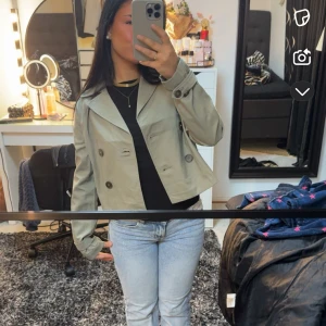 Grön croppad trenchcoat - Snygg croppad trenchcoat i beige med dubbelknäppning och stora knappar. Jackan har klassisk krage och långa ärmar med knappdetalj vid ärmslut. Perfekt för dig som vill ha en stilren och trendig look.