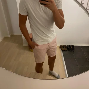 Rosa shorts från Tommy Hilfiger - Snygga ljusrosa shorts från Tommy Hilfiger med klassisk passform. De har fickor fram och bak, bälteshällor och en liten broderad logga bak. Tillverkade i mjuk bomull, perfekta för varma sommardagar. Stängs med knapp och dragkedja.