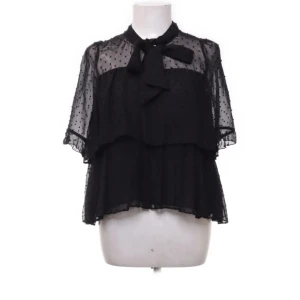 Svart prickig blus med rosett från Zara - Supersöt svart blus från Zara med transparenta partier och små prickar. Blusen har korta ärmar, volanger och en snygg rosett vid halsen. Perfekt för dig som gillar en luftig och trendig look med extra detaljer.