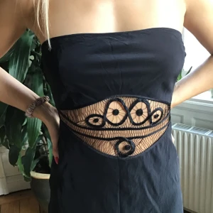 Svart axelbandslös långklänning - Svart axelbandslös långklänning med sensuell och unik meshdetalj , broderat mönster i midjan. Klänningen har en figurnära passform och öppen rygg, vilket ger en snygg och modern look. Perfekt för dig som vill fånga manliga blickar och  sticka ut med coola detaljer.