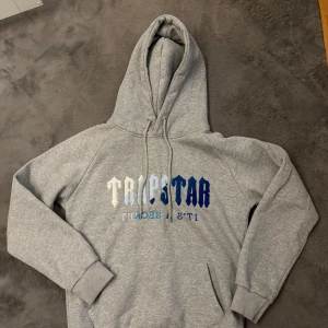 Grå Trapstar hoodie med blå text - Snygg grå hoodie från Trapstar med stor huva och snörning. Framsidan har Trapstar-logga i olika blå nyanser och vitt. Mjuk bomullskänsla, ribbade muddar och klassisk passform. Perfekt för dig som gillar streetwear och vill sticka ut lite extra. Använd fåtal ggr och i väldigt bra skick!! 