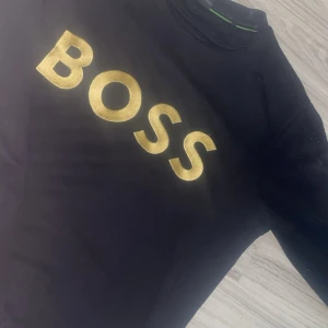 Svart BOSS t-shirt med guldtryck - Svart t-shirt från BOSS i storlek XS med stort, glansigt guldtryck över bröstet. Klassisk rund halsringning och korta ärmar. Perfekt för dig som vill sticka ut med en clean och lyxig vibe.