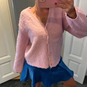 Ljusrosa fluffig kofta med knappar - Supermjuk och fluffig ljusrosa kofta med v-ringning och knäppning framtill. Koftan har lång ärm och en avslappnad passform, perfekt att slänga på sig över en topp. Enkel och söt design som passar till mycket.
