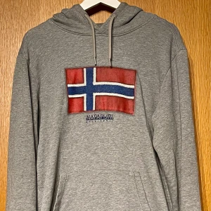 Napapijri Hoodie - Grå hoodie från Napapijri med stor norsk flagga på bröstet och logga under. Tröjan har huva med snörning, känguruficka och långa ärmar. Perfekt nu till kallare väder och ett riktigt kap i bra skick👌 Tveka inte på att höra av dig!
