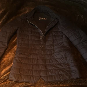 Herno Kavajjacka - Snygg svart quiltad jacka från Herno med klassisk kavajkrage, knappar och dragkedja. Jackan har två fickor framtill och diskret logga på ärmen. Perfekt för dig som vill ha en stilren och varm look under höst
