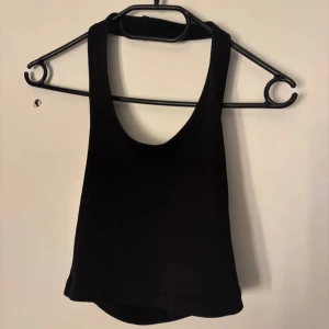 Svart ribbad croppad linnetopp - Säljer en svart ribbad linnetopp med croppad passform och djup urringning både fram och bak. Perfekt för dig som gillar en enkel men ändå edgy stil. Toppen är ärmlös och har en tight passning som framhäver siluetten.