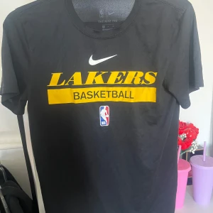 Svart Lakers t-shirt från Nike - Svart t-shirt från Nike med LA Lakers-tryck i gult och vitt samt NBA-logga på bröstet. Modellen är kortärmad och tillverkad i Dri-FIT-material som är tränings kläder material. Det finns inget tryck på baksidan eller något bara helt blankt 