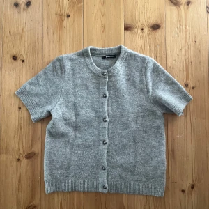 Grå stickad kofta från Gina Tricot - Kortärmad grå stickad kofta från Gina Tricot med rund hals och knappar framtill. Mjuk och skön, perfekt att slänga över en t-shirt eller linne. Ribbstickade muddar vid ärmslut och nederkant ger en clean look.