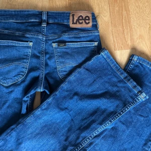 Blå lågmidjade Lee jeans  - Snygga blå lågmidjade bootcut jeans från Lee i modellen ”Jessica”, färgen ”last chapter”. Jeansen har fem fickor, låg midja och är gjorda i mjukt stretchigt denimtyg. Perfekta för dig som gillar en tidlös look med lite utsvängda ben. Stora Lee-loggan bak på linningen ger extra retrovibbar. I mycket fint skick. Storlek: W24 L31. Nypris:999kr. Säljer för:499kr.