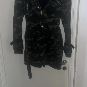 Camouflage parkajacka från Gothicana - Säljer en lång parkajacka i camouflage från Gothicana. Jackan har dragkedja, knappar, bälte i midjan och flera fickor. Insidan är fodrad med svart fleece för extra värme. Snygga detaljer som metallspännen och dragkedjor gör den unik. 