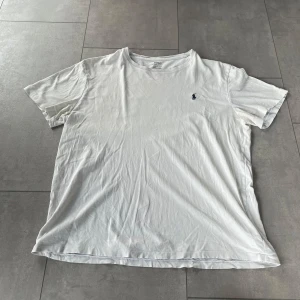 Vit t-shirt från Ralph Lauren - Säljer en klassisk vit t-shirt från Ralph Lauren med en liten broderad logga i blått på bröstet. T-shirten har en rund hals och korta ärmar, perfekt för en stilren look. Bra/okej skick. Trycket vid nacken är slitet men själva tröjan är bra