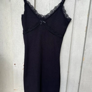 Svart spetsklänning från H&M Divided - Supersöt svart kortklänning från H&M Divided i ribbad bomull med spetsdetaljer vid halsringningen och en liten rosett framtill. Axelbandslös design som ger en snygg och enkel look, perfekt för dig som gillar stilrena vibes.
