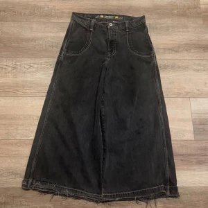 Svart lång jeanskjol från JNCO - Säljer en svart lång jeanskjol från JNCO med rå kant nertill och stora bakfickor. Ena bakfickan har broderad grön och gul graffiti-logga. Kjolen har klassiska jeanssömmar, framfickor och en avslappnad loose passform. Perfekt för dig som gillar streetstyle.