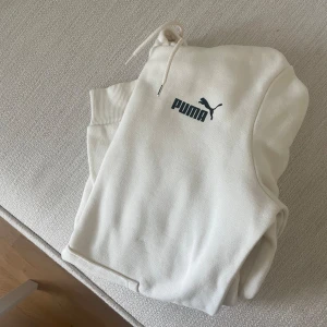 Vit hoodie från Puma med huva - Säljer en stilren vit hoodie från Puma med svart logga på bröstet. Tröjan har huva med snörning och ribbade muddar vid ärmsluten. Perfekt för en avslappnad och sportig look.