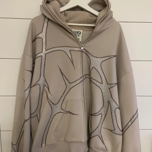 Named Collective Hoodie - En ljusbeige unisex hoodie med rhinestones från Named Collective! Dragkedjan funkar fint och kan öppnas/stängas både uppifrån och nerifrån. Den går även hela vägen till toppen av luvan. Storlek X/XL. Tyget är otroligt mjukt på insidan. Nästan som ny förutom att det tyvärr finns några fläckar på fickan. (Sista bilden) Hör gärna av dig om du har frågor!!