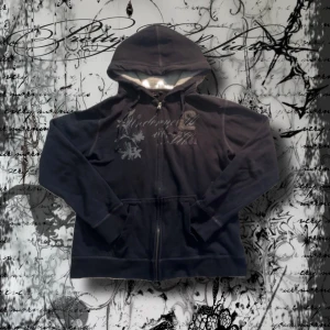 Svart hoodie med tryck från Equipe - Svart hoodie från Equipe med dragkedja och huva. Framsidan har coolt tryck med blommor, siffror och broderad logga. Mjuk insida och ribbade muddar. Perfekt för dig som gillar streetstyle och vill ha en chill look. Ingen tryck på baksidan. Width:52 length 56 