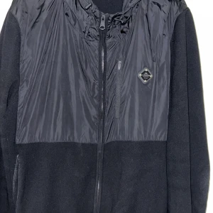 Svart vindjacka från J.Lindeberg - Säljer en svart vindjacka från J.Lindeberg med huva och dragkedja framtill. Jackan har en kombination av glansigt och matt material, samt en liten bröstficka med dragkedja och logomärke på bröstet. Perfekt för blåsiga dagar.