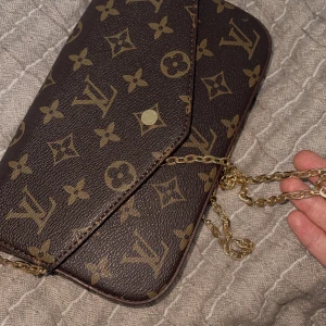 axelväska - Säljer en brun axelväska ،  klassiskt monogrammönster och guldfärgad kedja. Väskan har kuvertformad front med tryckknapp och vinrött innerfoder. Perfekt storlek för det viktigaste och ger en lyxig touch till din outfit.