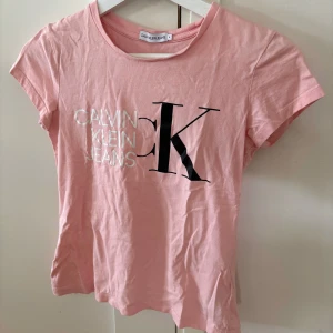 Rosa t-shirt från Calvin Klein Jeans - Snygg rosa t-shirt från Calvin Klein Jeans med stort CK-tryck i svart och vitt framtill. Modellen är kortärmad och har en klassisk rund halsringning. Perfekt för dig som gillar ett stilrent plagg och mjuk bomullskänsla. Storlek: 164 (passar som en xxs)