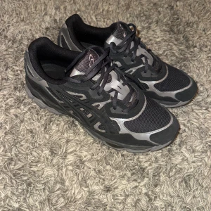 Asics Gel-NYC - Nypris 2000kr, säljer för 1000kr. Knappt använda. Säljer ett par Asics sneakers i svart och grått med coola detaljer och meshpaneler för extra andningsförmåga. Skorna har snörning och en chunky sula som ger skön komfort. Perfekta för dig som gillar sportig stil och vill ha något som sticker ut.