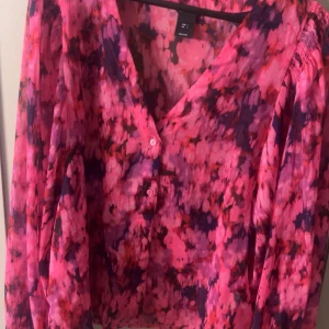 Blommig rosa blus från Lindex - Färgstark skjorta från Lindex i storlek L med blommigt mönster i rosa, lila och rött. Skjortan har v-ringning, knappar framtill och långa ärmar. Perfekt för dig som vill sticka ut med en cool och livlig look.