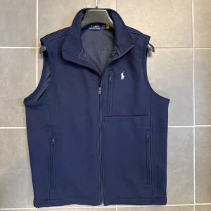 Marinblå väst från Polo Ralph Lauren - Snygg marinblå väst från Polo Ralph Lauren med hög krage och dragkedja framtill. Västen har meshfoder, tre fickor med dragkedja och klassisk vit logga på bröstet. Perfekt för lager-på-lager och stilren look.
