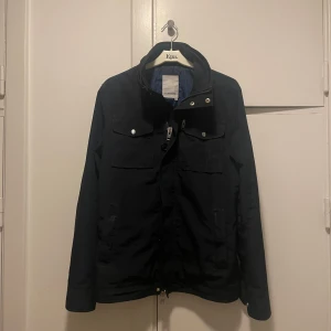 Svart vindjacka från J.Lindeberg - Svart vindjacka från J.Lindeberg med stilrena silverdetaljer och flera fickor framtill. Jackan har hög krage, dragkedja och tryckknappar. Perfekt för dig som gillar en clean och sportig look. Klassisk passform och diskret logga på ärmen.