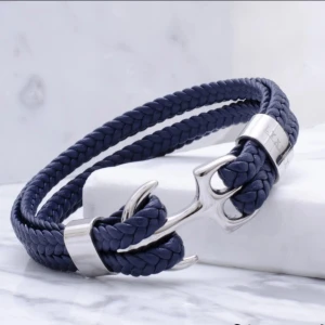 Blått armband med ankardetalj - Snyggt armband i flätat blått material med en cool silverfärgad ankare som lås. Perfekt accessoar för dig som gillar marina vibbar och stilrena detaljer. Armbandet har även silverfärgade metalldetaljer som ger en extra touch.