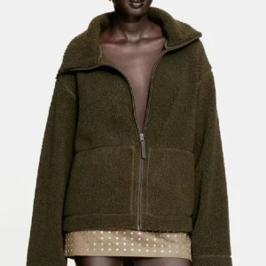 Olivgrön teddyjacka med dragkedja - Mysig olivgrön teddyjacka med oversized passform och hög krage. Jackan har en grov dragkedja framtill och är tillverkad i fluffigt teddy-material som ger en cool och avslappnad vibe. Perfekt för lager-på-lager-stil under kyliga dagar.