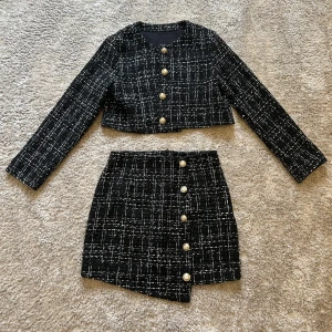 Svart rutig set med guldknappar - Supersnygg svart set med vit rutigt mönster och fem dekorativa guldknappar framtill. Kjolen har en lätt asymmetrisk nederkant och är gjord i ett tjockare, strukturerat material som ger en lyxig känsla. Perfekt att matcha med en croppad jacka för en trendig look. Köpt i Sinsay. 