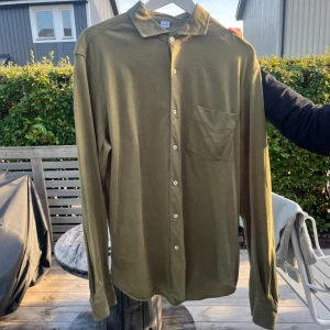 Olivgrön skjorta från Aspesi - Snygg olivgrön skjorta från Aspesi i bomull med klassisk krage, vita knappar och bröstficka. Skjortan har lång ärm och en clean, stilren look som passar perfekt till jeans eller chinos. Köpt för   1400, som ny