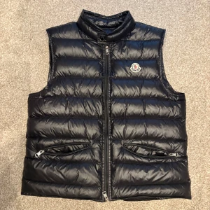 Svart dunväst från Moncler - Snygg svart dunväst från Moncler med klassisk quiltad design och hög krage. Västen har två fickor med dragkedja och ikonisk Moncler-logga på bröstet. Perfekt att slänga över en hoodie för en clean och sportig look.