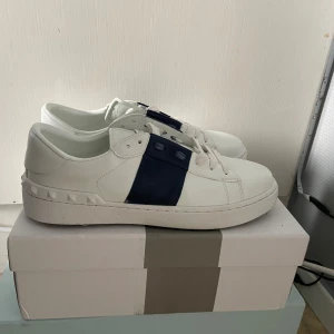Valentino Garavani opens - Snygga vita sneakers från Valentino Garavani med en bred mörkblå rem över mitten och coola nitar längs hälen. Skorna har rund tå, platt sula och är gjorda i skinn. Perfekta för dig som vill ha en stilren men ändå unik look. Ställ gärna frågor angående skorna! Priset kan diskuteras 