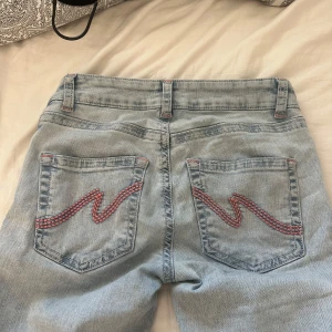 Bootcut jeans från Nelly med slitningar - Ljusblå bootcut jeans från Nelly med låg midja och coola slitningar vid knäna. Bakfickorna har rosa broderade detaljer som ger extra edge. Klassisk femficksmodell i mjuk bomullsdenim med dragkedja och knapp framtill.